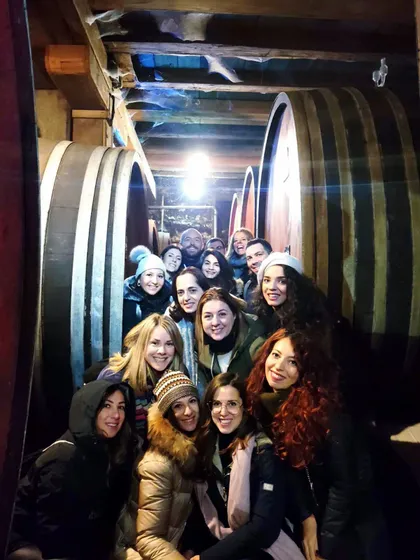 Eine WeRoad-Gruppe lachender Menschen, die für ein Foto in einem engen Weinkeller zwischen großen Holzfässern posieren.