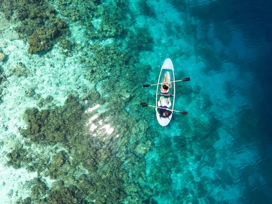 Una vista aerea di due persone che remano in un kayak trasparente sopra una barriera corallina in acqua turchese limpida.