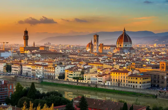 A historic cityscape featuring a large dome and a tower, illuminated by the golden light of sunset, with a river in the foreground.