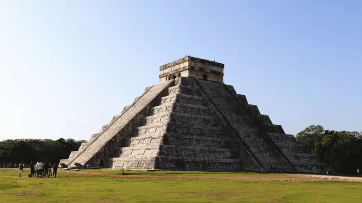 Un gruppo WeRoad in viaggio si trova su un campo erboso, ammirando una grande piramide a gradoni di pietra sotto un cielo azzurro e limpido.