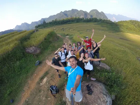 Eine WeRoad-Gruppenreise macht ein lächelndes Selfie auf einem Wanderweg mit Blick auf terrassierte grüne Hügel und ferne Berge.