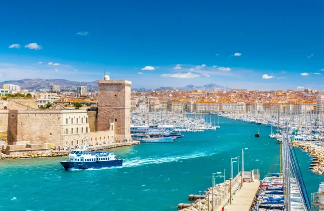 Un traghetto naviga su acque turchesi oltre una fortezza di pietra all'ingresso di un porto pieno di barche, con uno skyline della città sullo sfondo.
