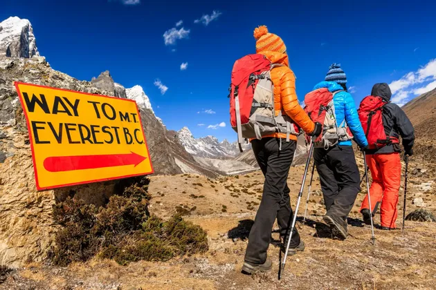 Eine WeRoad-Gruppe von drei Wanderern mit Rucksäcken und Stöcken wandert auf einem Bergpfad an einem gelben Wegweiser vorbei.