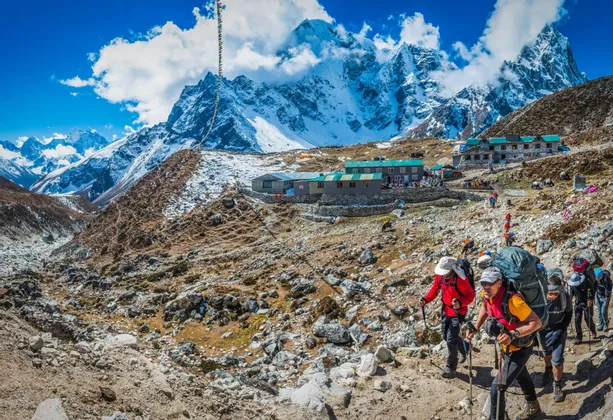 Eine WeRoad-Gruppenreise, die auf einem felsigen Pfad zu einer Berghütte wandert, mit großen, schneebedeckten Gipfeln im Hintergrund.