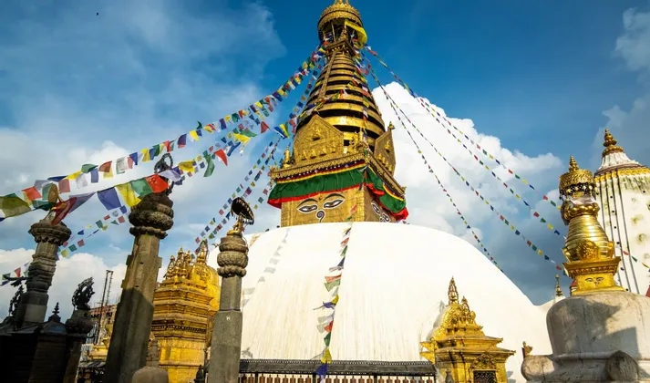 Eine Aufnahme aus der Froschperspektive zeigt eine große, weiße Stupa mit goldener Spitze und gemalten Augen, geschmückt mit bunten Gebetsfahnen, vor blauem Himmel.