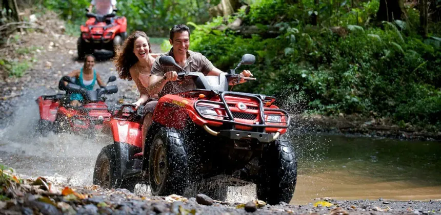 Un gruppo WeRoad sorride mentre attraversa un ruscello poco profondo su ATV rossi in una giungla lussureggiante, schizzando acqua.