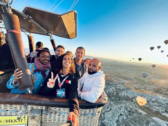Un groupe WeRoad prend un selfie dans la nacelle d'une montgolfière, volant haut au-dessus d'un paysage parsemé d'autres ballons au lever du soleil.