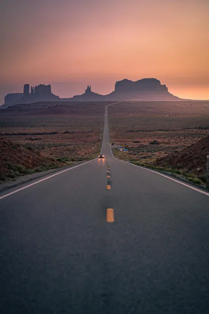 Un'auto solitaria con i fari accesi percorre una strada dritta nel deserto verso mesas lontane sotto un cielo rosa e tenue al crepuscolo.
