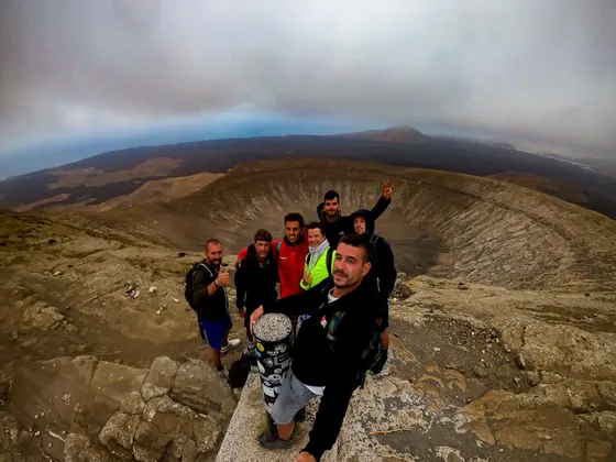 Eine WeRoad-Gruppe posiert für ein Selfie am felsigen Rand eines Vulkankraters, mit einer weiten Landschaft darunter.