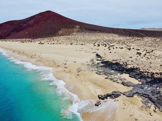 Eine Luftaufnahme eines Sandstrandes mit türkisfarbenem Wasser und vulkanischen Felsen, mit einem großen, roten Berg im Hintergrund.