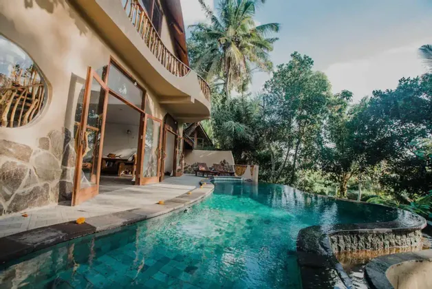 Una piscina a sfioro con acqua turchese accanto a una villa moderna con porte a vetro aperte, circondata da una rigogliosa foresta tropicale.