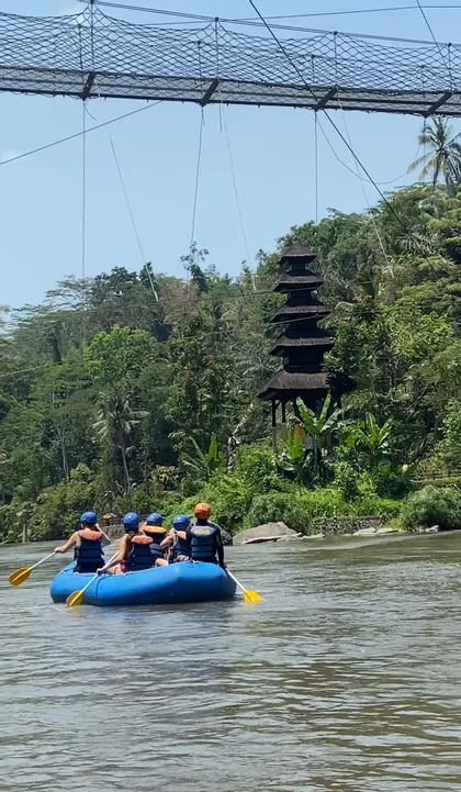 Un viaggio di gruppo WeRoad in rafting su una barca blu in un fiume, passando un tempio e una giungla lussureggiante sotto un ponte sospeso.
