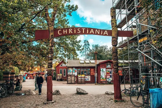 Der Eingang zu Christiania, gekennzeichnet durch ein Holzschild und zwei große geschnitzte Säulen, an einer Kopfsteinpflasterstraße mit Bäumen.