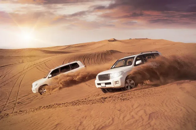 Deux SUV blancs descendent une grande dune de sable dans le désert, soulevant des nuages de sable sous un soleil couchant.