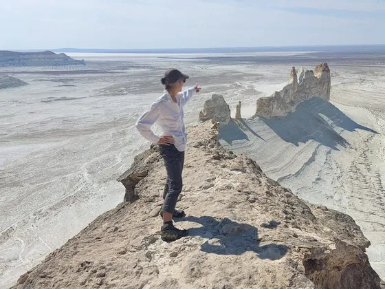 Una persona con un cappello si erge su una stretta cresta rocciosa, indicando una vasta valle desertica illuminata dal sole con formazioni rocciose bianche.