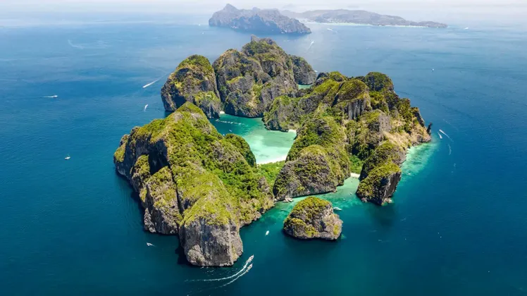 Vue aérienne d'un archipel rocheux et verdoyant dans un océan d'un bleu profond, avec des bateaux naviguant autour d'un lagon central turquoise.