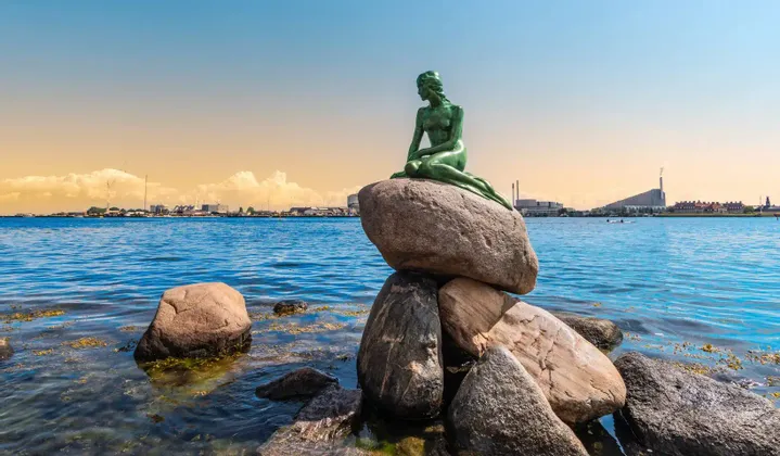 Eine Bronzestatue der Kleinen Meerjungfrau sitzt auf einem Felsen am Meer, mit einer fernen Stadtkulisse am Horizont unter klarem Himmel.
