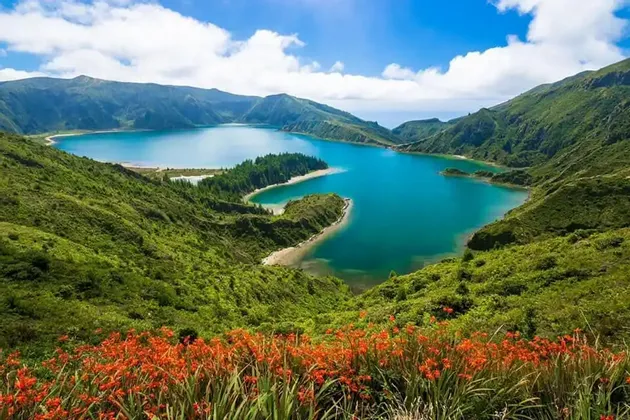 Una vista panoramica da una collina con fiori arancioni in primo piano, che si affaccia su un lago vulcanico di un blu brillante circondato da montagne verdi.