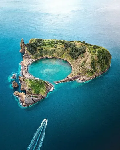 Vista aerea di un isolotto vulcanico verde con una laguna centrale turchese, mentre una motobarca passa nel mare blu profondo.