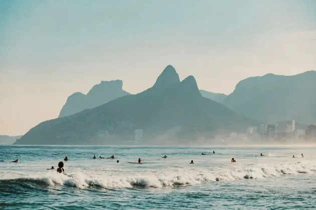 Surfisti in attesa delle onde nell'oceano con grandi montagne e uno skyline cittadino sullo sfondo.