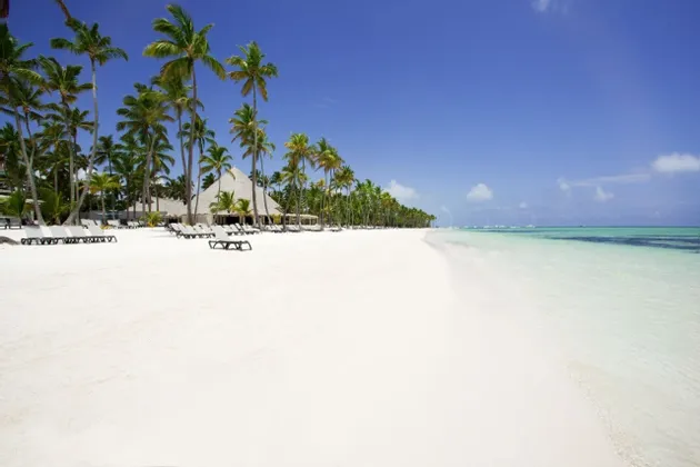 Un'ampia spiaggia di sabbia bianca fiancheggiata da palme e sedie a sdraio accanto a un'acqua cristallina color turchese.