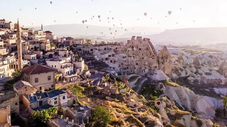 Una vista amplia de un pueblo tallado en colinas rocosas con numerosos globos aerostáticos flotando en el cielo al amanecer.