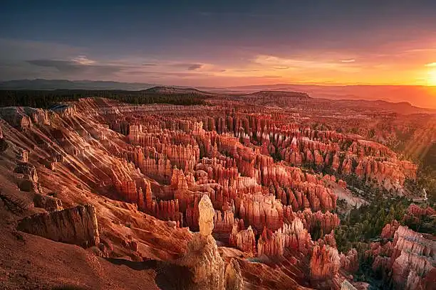 Un vasto canyon, pieno di camini delle fate e guglie di roccia rossa, illuminato dalla calda luce di un tramonto, con una foresta sullo sfondo.