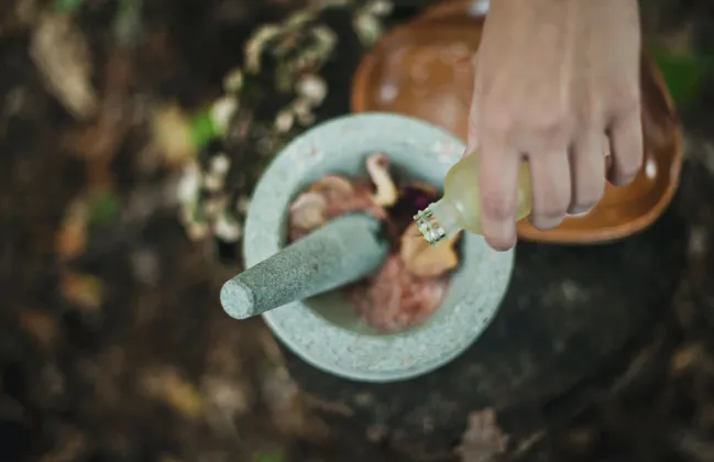 Una vista dall'alto di una mano che versa un liquido da una bottiglia in un mortaio e pestello di pietra con ingredienti naturali all'interno.