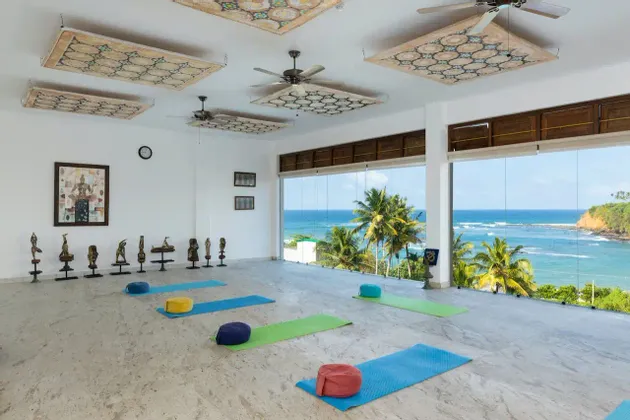 Uno studio yoga luminoso e vuoto con tappetini e cuscini su un pavimento di marmo, con ampie finestre che mostrano un oceano tropicale e palme.