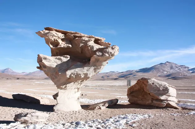 Una gran formación rocosa erosionada por el viento se alza en un desierto arenoso con montañas nevadas a lo lejos bajo un cielo azul claro.